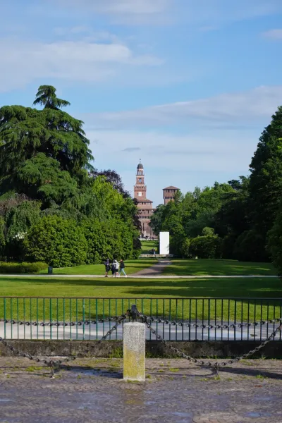 La torre del Castello Sforzesco si erge sopra gli alberi nel Parco Sempione, Milano, con persone che camminano su un sentiero in primo piano.