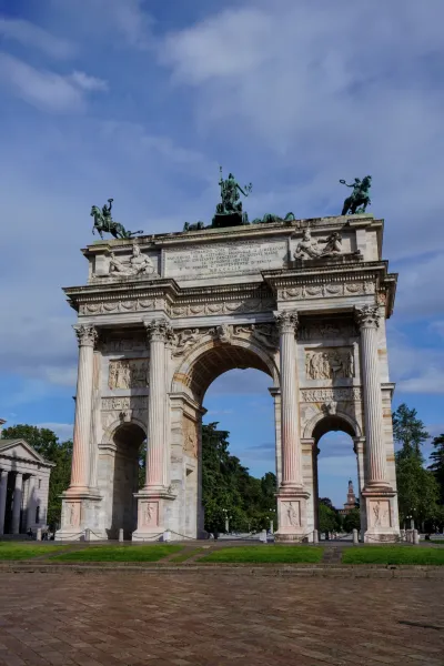 L'Arco della Pace, un grande arco trionfale neoclassico, si erge contro un cielo blu parzialmente nuvoloso, con alberi e erba in primo piano.
