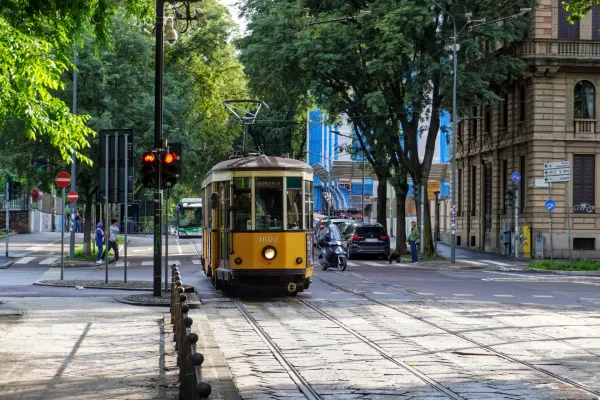Eine gelbe Vintage-Straßenbahn mit der Nummer 1602 fährt auf Gleisen auf einer Straße in Mailand, mit Bäumen, Gebäuden, einem Bus, einem Auto und einem Roller im Hintergrund.