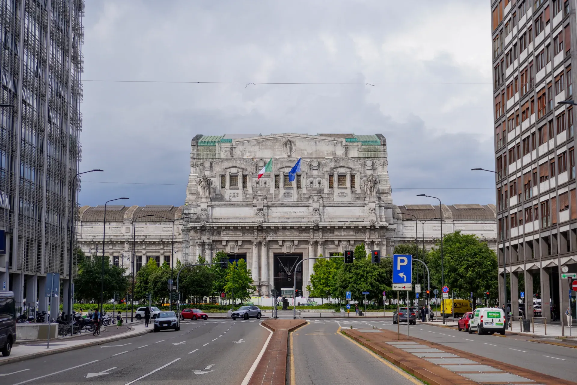 L'impressionante facciata della Stazione Centrale di Milano con bandiere italiane ed europee, circondata da alberi e con traffico sulla strada in primo piano.