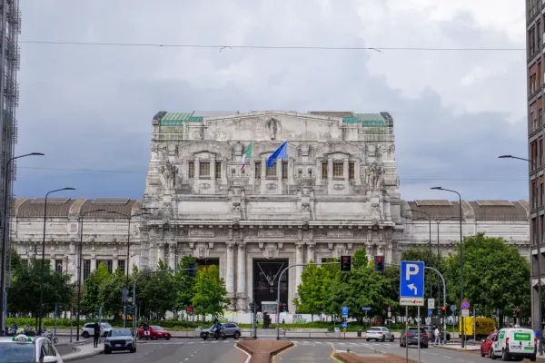 L'imponente facciata della Stazione Centrale di Milano con bandiere italiane ed europee in una giornata nuvolosa, con auto e alberi sulla strada in primo piano.