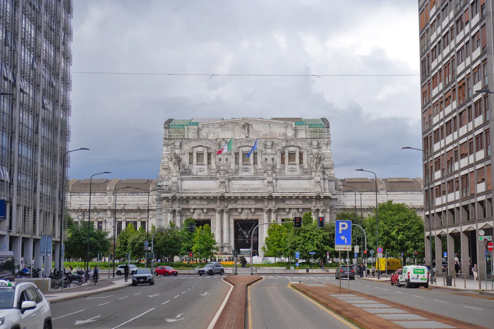 Pogled na Centralnu Stanicu u Milanu sa njenom složenom fasadom, zastavama, modernim zgradama, ulicom, automobilima i drvećem pod oblačnim nebom.