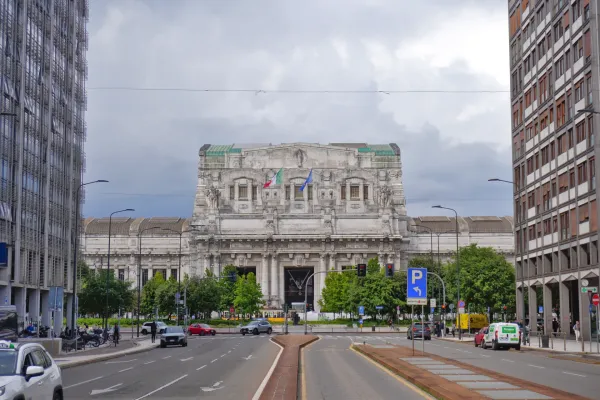 Vista della Stazione Centrale di Milano con la sua elaborata facciata, bandiere, edifici moderni, strada, auto e alberi sotto un cielo nuvoloso.