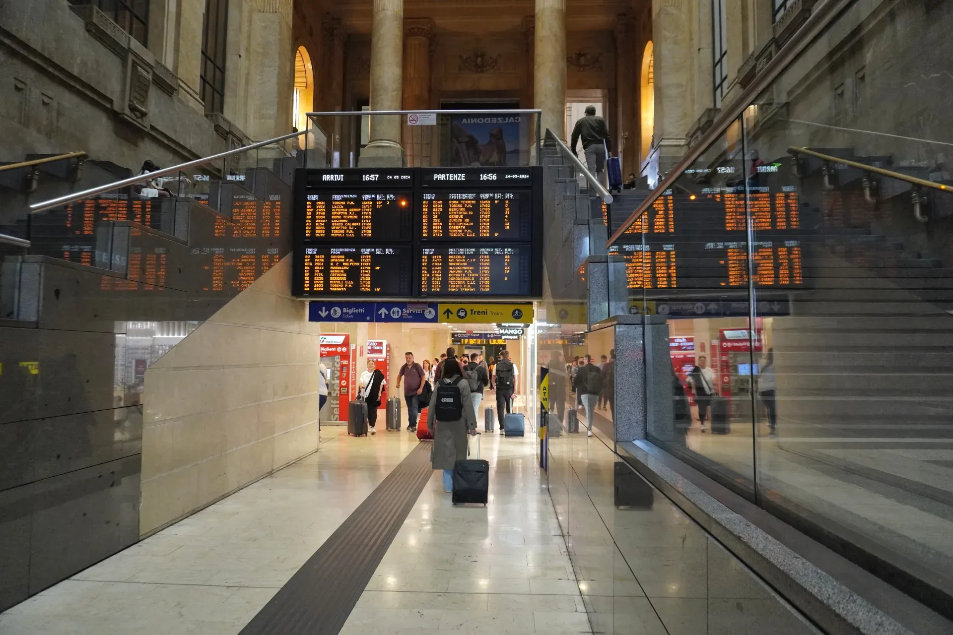 Reisende mit Koffern in der großen Halle des Bahnhofs Milano Centrale, mit elektronischen Abfahrts- und Ankunftstafeln, die Zuginformationen anzeigen.